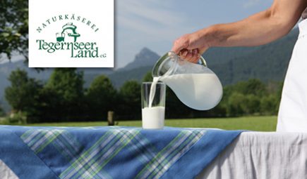 Logo Naturkäserei Tegernseer Land. Tisch mit Glas, in das Milch eingeschenkt wird.